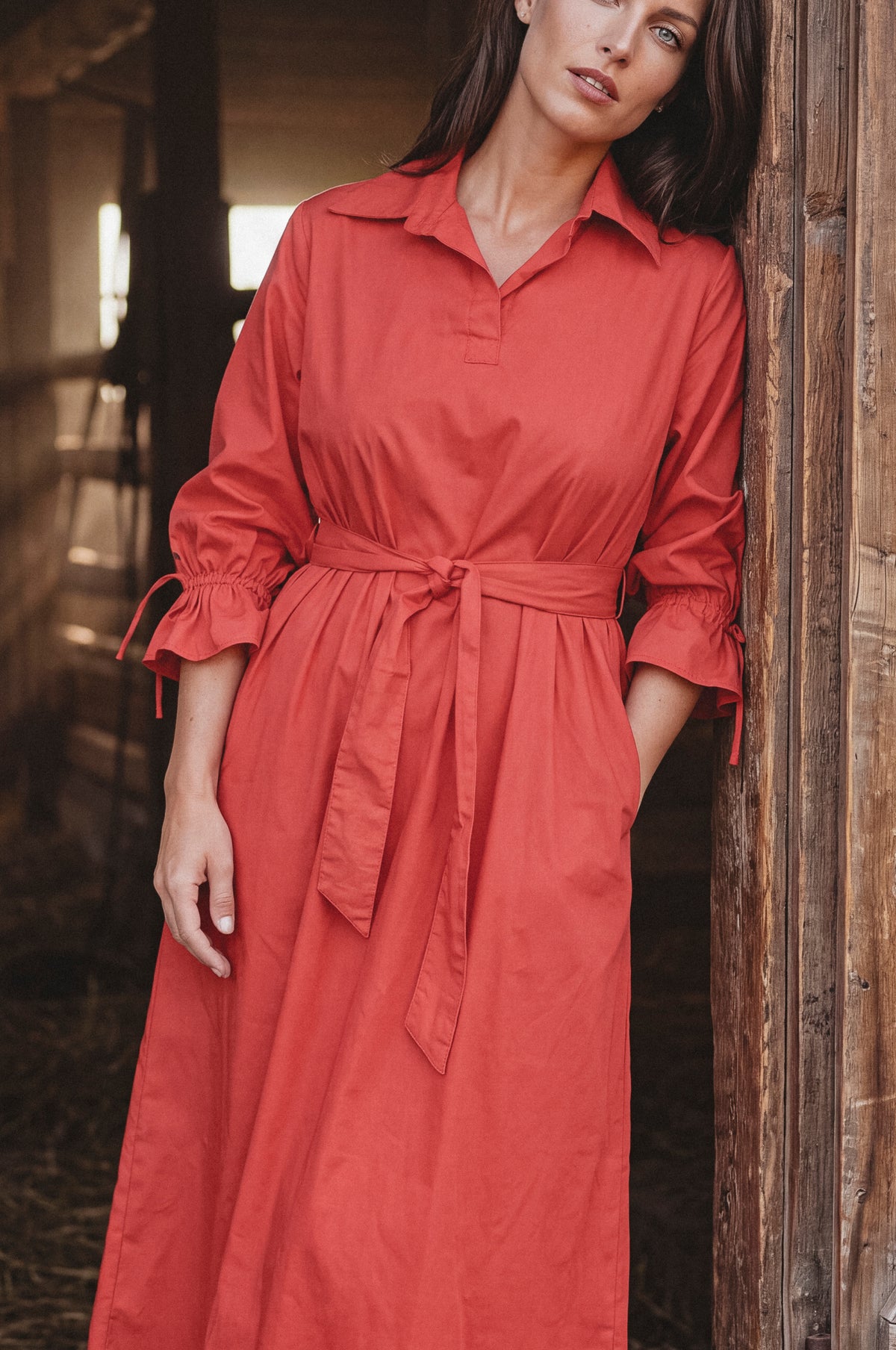A woman wearing a rust-red long-sleeve collared shirt dress with a matching waist tie, standing in a rustic barn next to a wooden post.