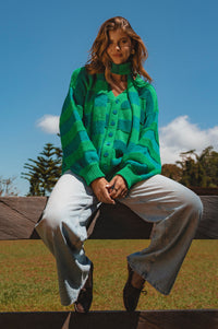 Person wearing a green sweater and light pants sitting on a wooden bench outdoors.