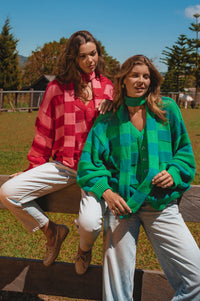 Two women sitting on a wooden bench outdoors, one wearing a red checkered shirt and the other in a green plaid sweater.
