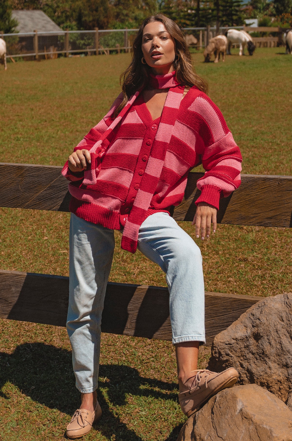 Woman wearing a red and pink checkered cardigan sitting on a wooden fence with a pastoral background.