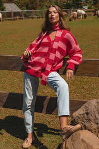 Woman wearing a red and pink checkered cardigan sitting on a wooden fence with a pastoral background.