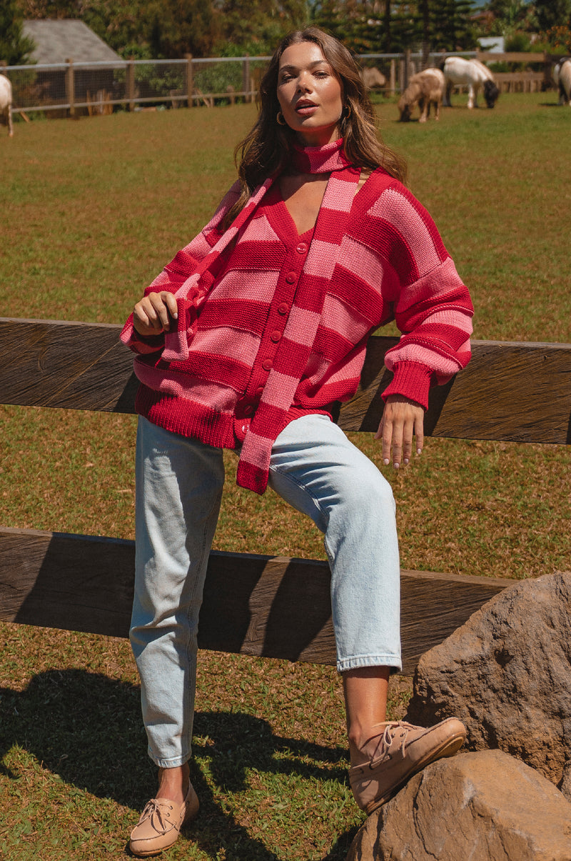 Woman wearing a red and pink checkered cardigan sitting on a wooden fence with a pastoral background.