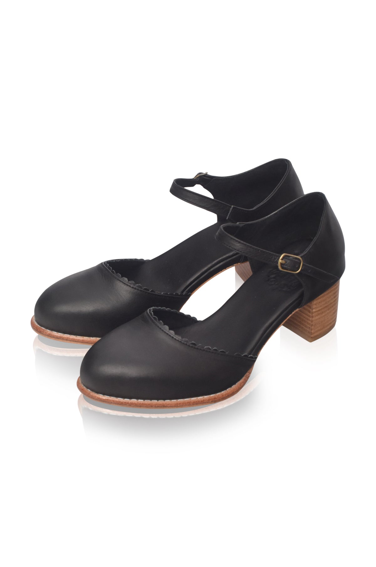 A pair of black leather Mary Jane style shoes with low block heels, ankle straps, and a small gold buckle, shown on a white background with a soft reflection.