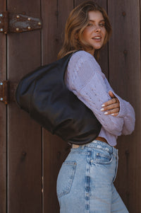 Woman wearing a black leather jacket and light purple sweater against a wooden background