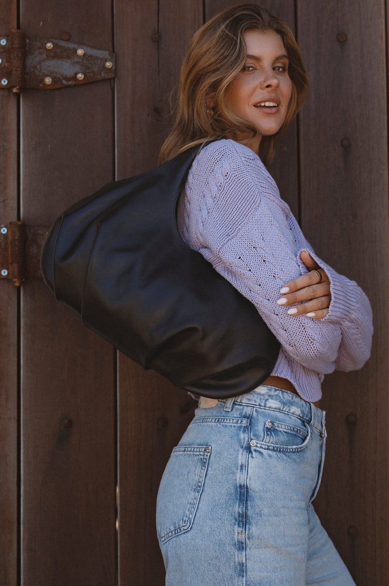 Woman wearing a black leather jacket and light purple sweater against a wooden background