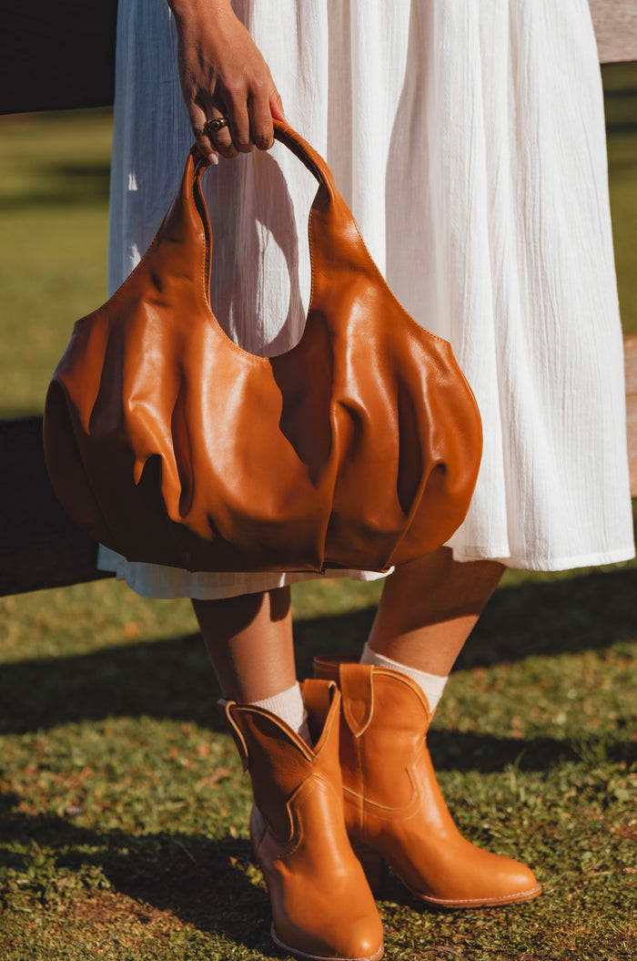 Person wearing a white dress and brown leather boots, holding a matching brown leather handbag on grass.