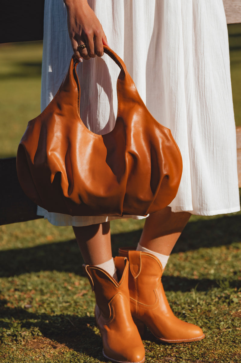 Person wearing a white dress and brown leather boots, holding a matching brown leather handbag on grass.