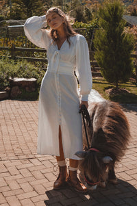 Woman in a white dress standing with a small brown pony on a brick path.