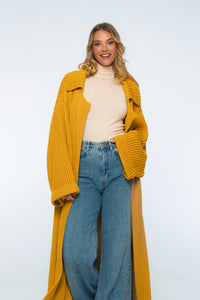 Woman wearing a mustard yellow cardigan, beige turtleneck, and blue jeans on a white background