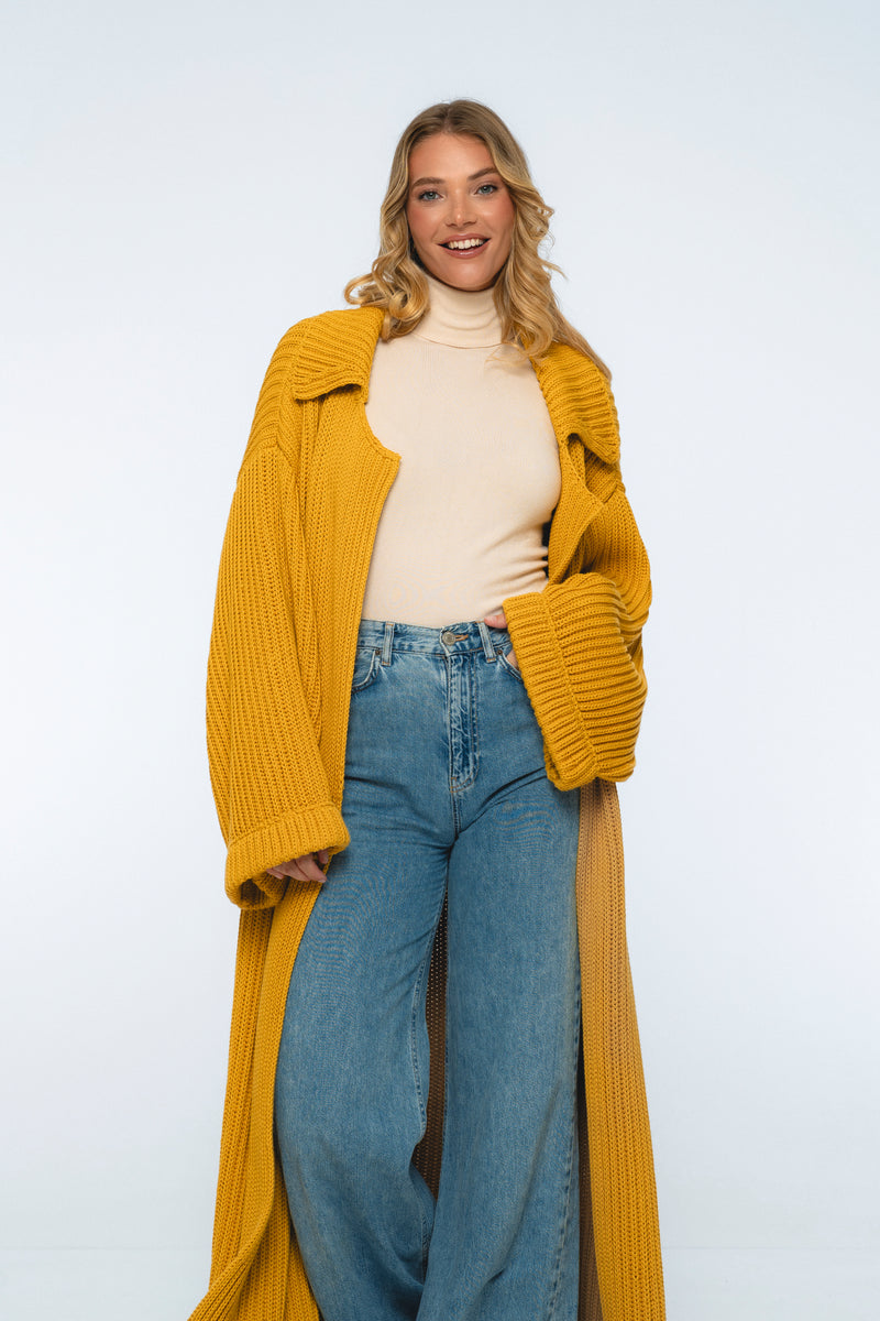 Woman wearing a mustard yellow cardigan, beige turtleneck, and blue jeans on a white background