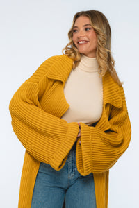 Woman wearing a mustard yellow cardigan over a beige top and blue jeans on a white background
