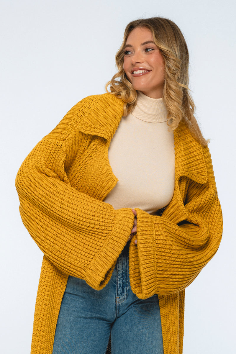 Woman wearing a mustard yellow cardigan over a beige top and blue jeans on a white background