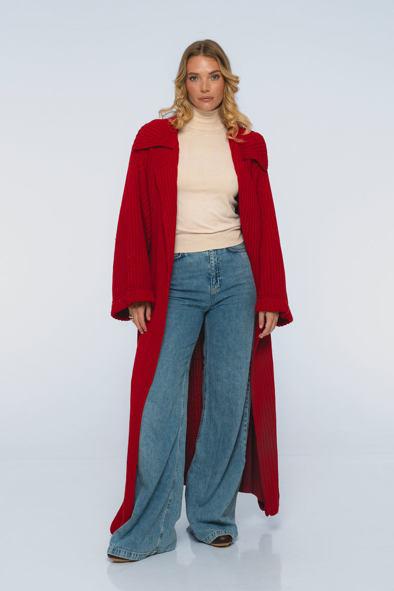 Woman wearing a long red coat, beige sweater, and blue jeans on a white background