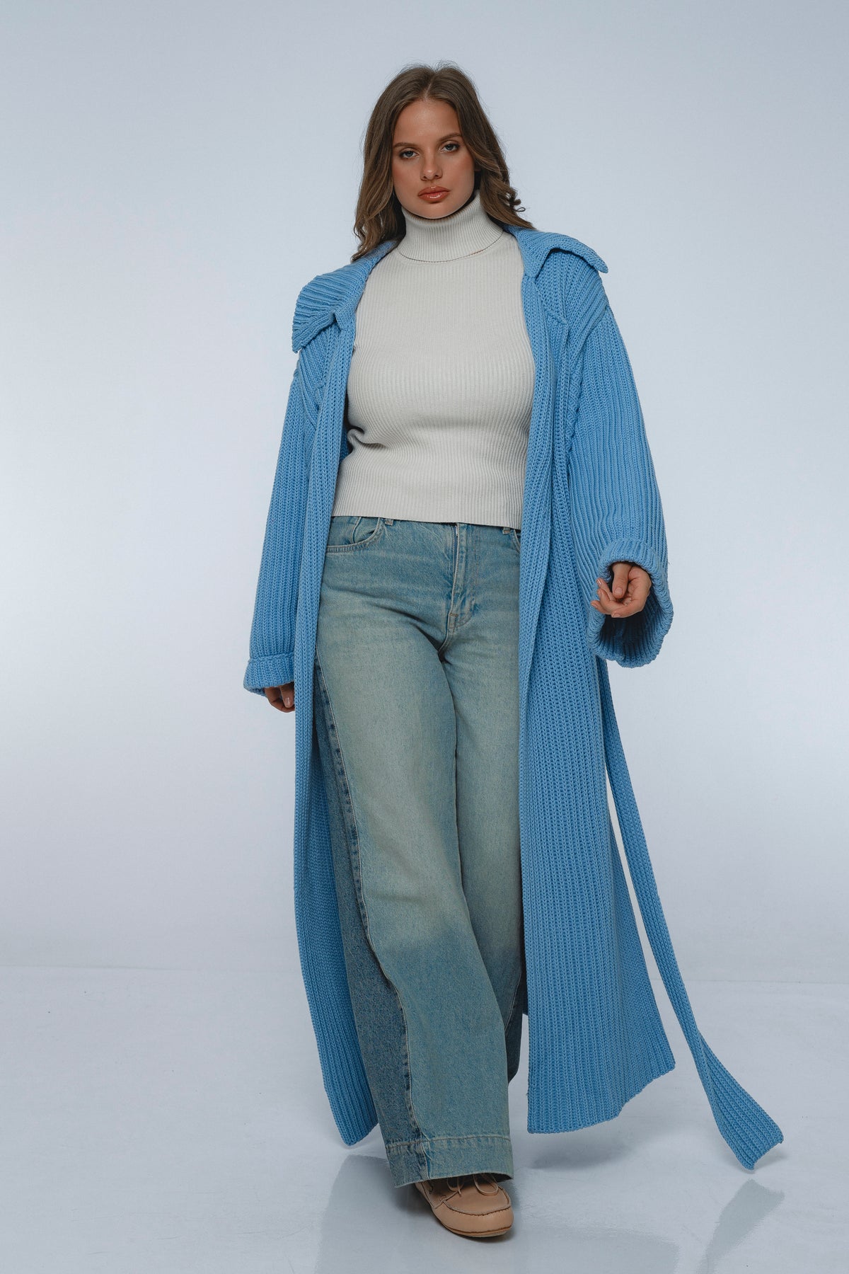 Woman wearing a long blue coat over a white turtleneck and jeans on a white background