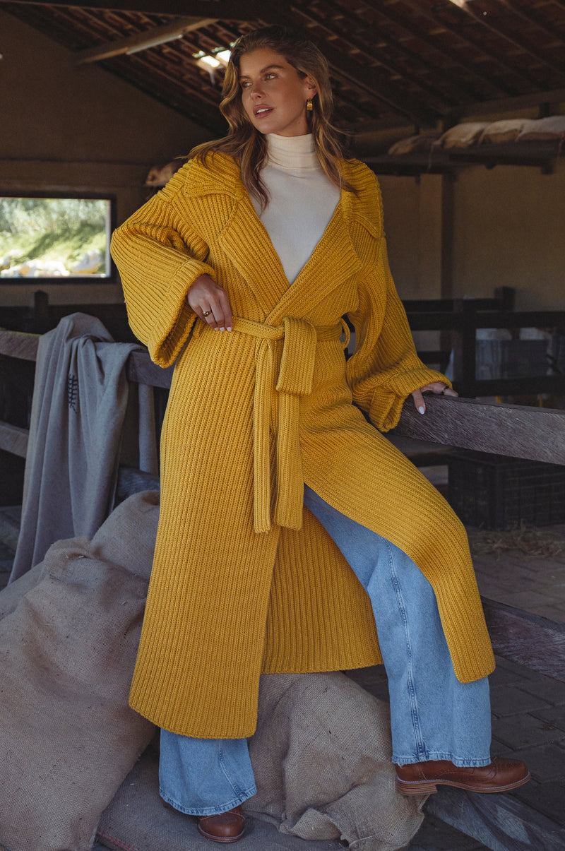 Woman wearing a yellow knitted coat sitting on a wooden bench in the barn.