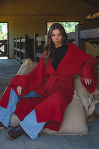 Woman wearing a red sweater and blue jeans sitting on a horse stall.