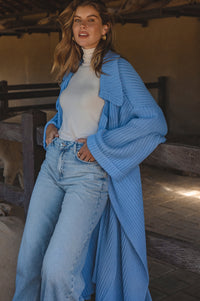 Woman wearing a blue knitted cardigan over a white top and jeans, standing in a rustic indoor setting.